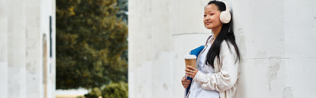 A young student with headphones leans against a wall, savoring coffee and embracing campus life.の写真素材