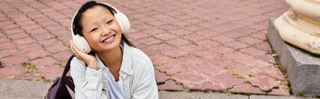 A stylish young girl with headphones smiles while lounging on the campus path, embracing student life.の写真素材