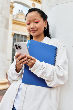 A young student with a trendy outfit engages with her smartphone near the university campus, displaying confidence.の写真素材