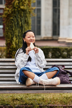 A stylish young woman relaxes on a park bench, savoring a snack, embodying youth culture at her university.の写真素材