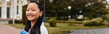 A young Asian student is smiling brightly while holding books near her university on a vibrant day.の写真素材