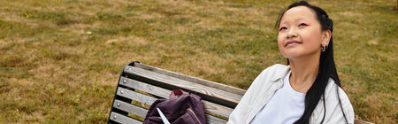 A pretty young Asian woman sits on a bench, smiling and reflecting while studying on her university campus.の写真素材