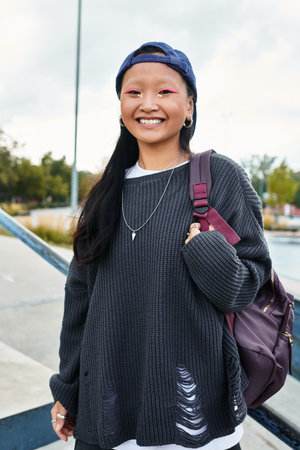 A cheerful young Asian girl in a trendy outfit stands outdoors, radiating joy and style.