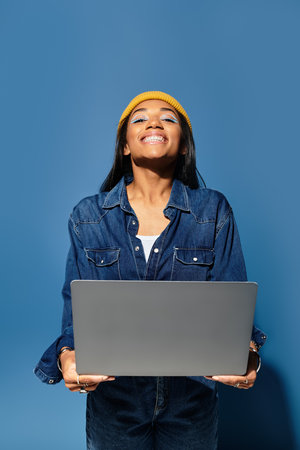 Smiling young woman in a cozy outfit stands with a laptop against a blue backgroundの写真素材
