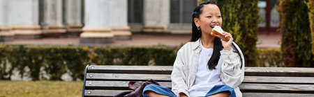 A cheerful young Asian student in a fashionable outfit eating sandwich while relaxing on a bench.の写真素材