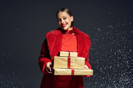 A cheerful woman in festive red beams while carrying wrapped presents in winter.の写真素材