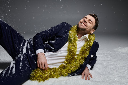 A joyful young man in formal wear lounges in fluffy white snow, adorned with festive decorations.