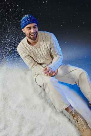 A cheerful young man sits on snow, enjoying the winter vibes while smiling in soft clothing.の写真素材