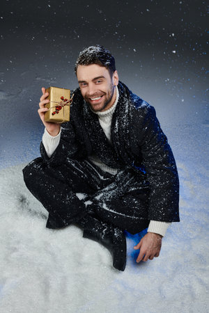 A cheerful young man dressed in winter attire sits in snow, holding a special gift with happiness.の写真素材