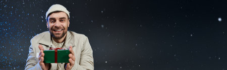 Smiling young man in a cozy outfit holds a present while enjoying the winter atmosphereの写真素材