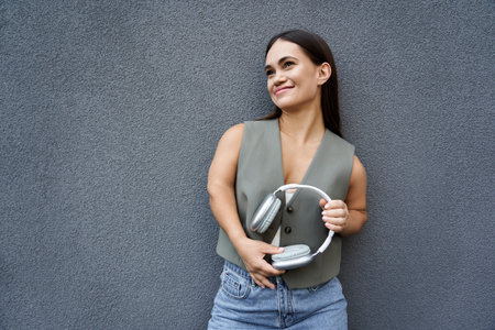 A young brunette woman with a cheerful smile takes a moment outside, holding her headphones while enjoying the fresh air.の写真素材