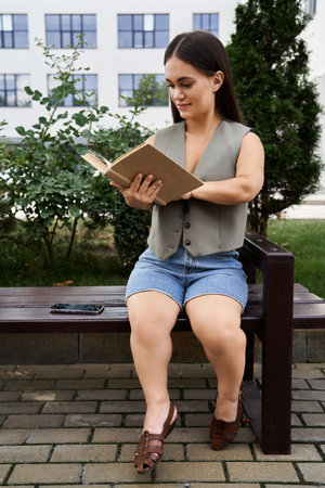 A young brunette woman with a charming smile is immersed in a book, savoring a tranquil outdoor moment surrounded by greenery.の写真素材