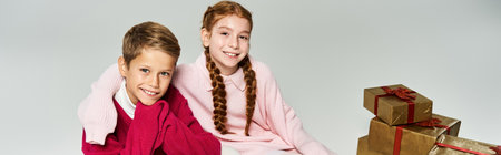 Two joyful kids in warm winter clothing are posing with holiday presents nearby.の写真素材