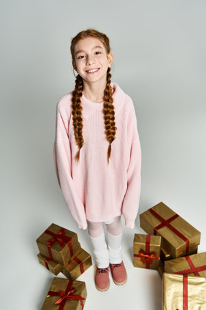 Delighted young girl wearing a pink sweater stands with festive presents, capturing holiday cheer.
