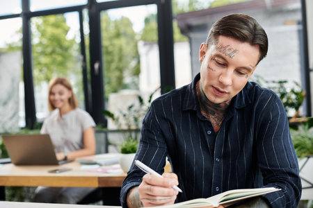 A focused young man writes in a notebook, while a smiling woman works at her desk.の写真素材
