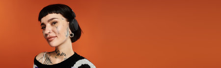 A confident young woman stands against a bright orange backdrop, flaunting her tattoos and colorful sweater.の写真素材