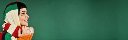 A cheerful young woman with tattoos and short hair smiles while wrapped in a cozy sweater and scarf, surrounded by green.の写真素材