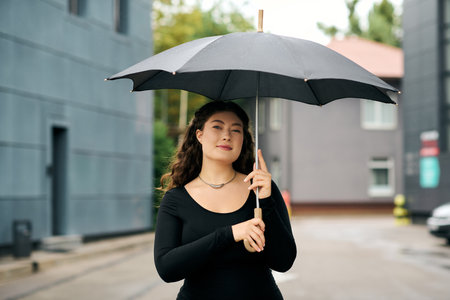 A stylish plus-size woman strolls through a quaint neighborhood, holding an umbrella on an autumn day.の写真素材