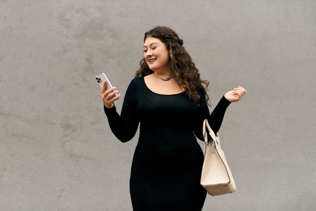 A young plus size woman gracefully walks outdoors on a crisp autumn day, checking her phone while...