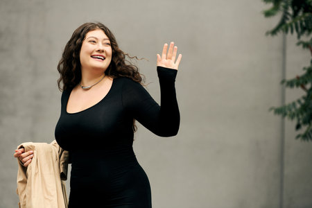 Brightly smiling, a young woman in a black dress waves warmly while enjoying the autumn air...