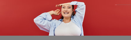 A young plus size woman showcases her vibrant makeup while posing joyfully in a bold red studio setting.の写真素材