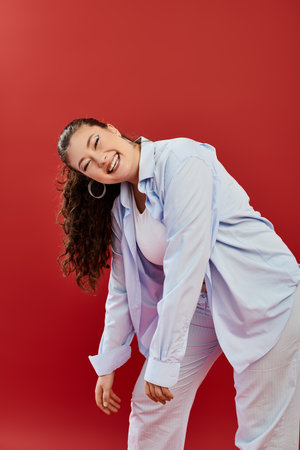 A joyful plus size woman showcases her bright makeup while posing playfully in a bold red studio...