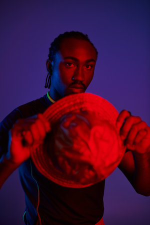 A young man showcases his fashion sense, holding a unique hat against a backdrop of glowing neon...