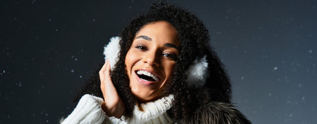 A cheerful, stylish brunette woman in earmuffs and a fur jacket smiles warmly in a winter-themed...