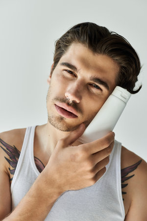 A stylish young man in a tank top leans against a hair product, showcasing his tattoos on a grey...