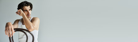 A tattooed young man in a tank top leans thoughtfully on a chair, lost in his own reflections.の写真素材