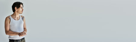 A young, handsome man in a tank top stands thoughtfully, showcasing his tattoos against a grey backdrop.の写真素材