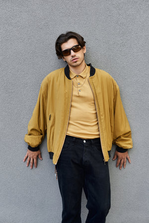 A young man showcases his fashionable autumn outfit while leaning casually against a gray wall.