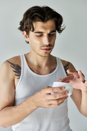 A young man in a tank top applies skincare cream, showing off his tattoo against a grey backdrop.の写真素材