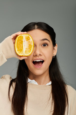 Emotional young woman in stylish attire playfully surprises with a slice of orange, expressing joy.の写真素材