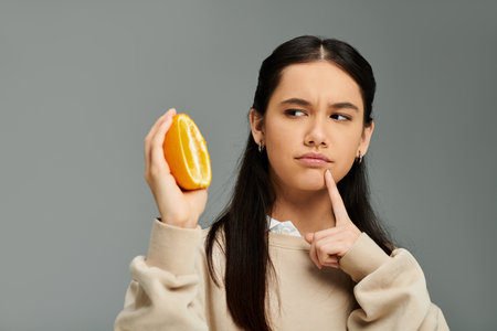A young woman in stylish attire thoughtfully examines a halved citrus fruit while posing.の写真素材