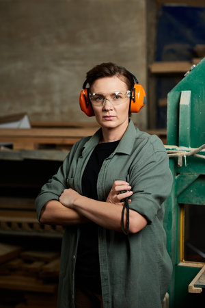 A skilled carpenter stands focused with safety gear on, surrounded by wood and tools in her...