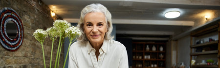 A joyful mature woman stands proudly with fresh flowers, radiating warmth and charm.の写真素材