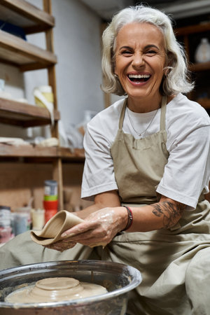 A mature woman smiles brightly while shaping clay at her pottery wheel, surrounded by creativity.の写真素材