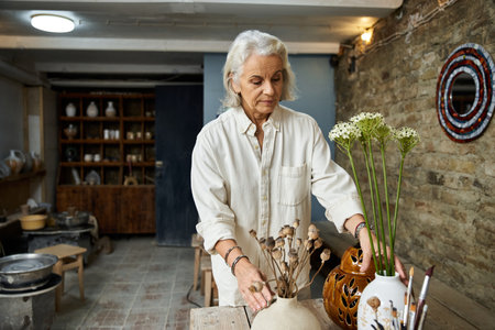 A beautiful mature woman carefully arranges delicate flowers in vases on a rustic wooden table.の写真素材
