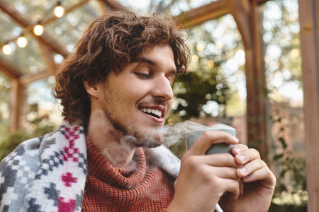 Enjoying a hot beverage while wrapped in a cozy blanket, a young man smiles brightly amidst a lush garden setting. Soft morning light filters through foliage, creating a tranquil atmosphere.の写真素材