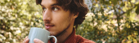 A charming young man with wavy hair sips from a mug, basking in the beauty of nature. The sunny day highlights the lush surroundings, creating a serene atmosphere.の写真素材