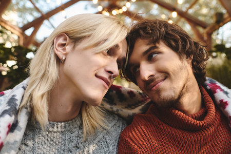Two handsome young men share a tender moment together, wrapped in a warm blanket in a beautiful outdoor space adorned with soft lights. Their joyful expressions reflect a deep bond and affection.の写真素材