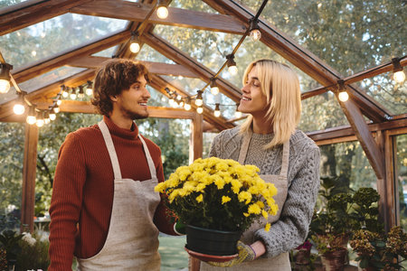 Brightly lit greenhouse filled with vibrant plants. A young, handsome couple engages in joyful conversation while admiring a pot of cheerful yellow flowers. They share smiles under warm string lights.の写真素材