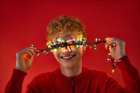 In a vibrant red setting, a young man beams with joy, playfully holding colorful Christmas lights across his eyes.の写真素材