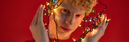 A confident young man flaunts trendy holiday fashion while playfully draping colorful Christmas lights around himself. Set against a vibrant red background, he exudes cheerful holiday spirit.の写真素材