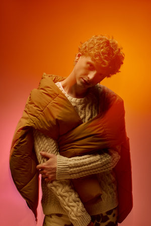 A stylish man showcases a creative holiday outfit, blending cozy textures with bold colors. He confidently wears a knitted sweater and a draped puffer, embodying contemporary Christmas vibes in 2025.の写真素材