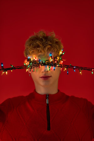 Brightly lit Christmas lights drape across a mans face, embodying the holiday spirit in a striking red setting. His joyful expression showcases his stylish seasonal outfit, perfect for celebrations.の写真素材