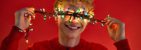 A cheerful young man dressed in festive red attire playfully holds colorful Christmas lights in front of his face. His bright smile radiates holiday spirit against a striking red background.の写真素材