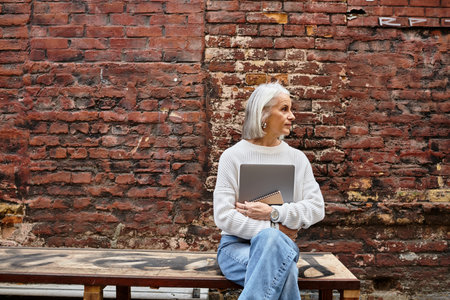 An elegant woman with gray hair contemplates life while seated on a rustic bench in an urban alley.の写真素材