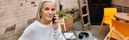 A beautiful gray haired woman sits at a table, engaged in a lively phone conversation outdoors.